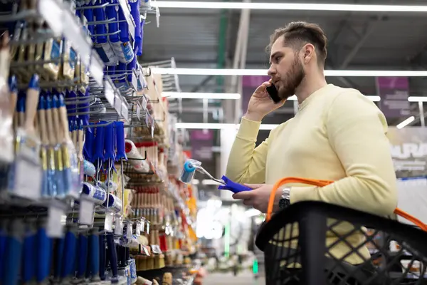 a man in a hardware store chooses paint brushes while discussing it on the phone with his wife.