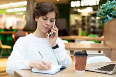 Esmer, bir kafede dizüstü bilgisayarda çalışıyor. Telefonda konuşuyor. Not defterine bir şeyler yazıyor..