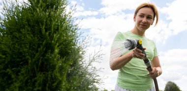 Genç kadın bahçıvan, çimlere hortumla su dökerek bakıyor..