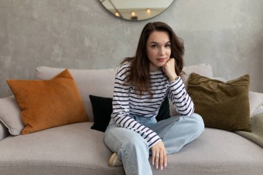 Kanepede oturan genç bir kadın eline yaslanmış kameraya bakıyor..