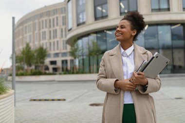 Genç tatmin olmuş Afrikalı Amerikalı bir kadın dizüstü bilgisayarla şehirde dolaşıyor ve gülümsüyor..