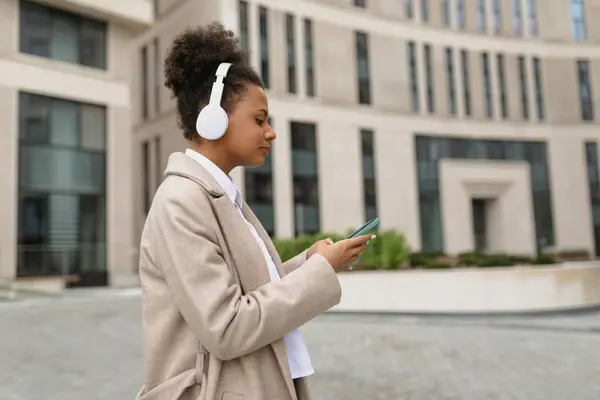 young african american woman hurrying to work listening to music in headphones via mobile phone against the backdrop of an office building.