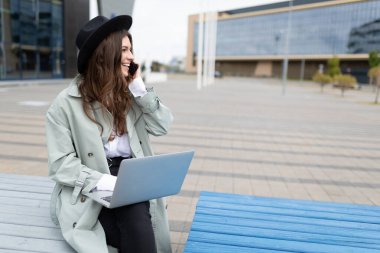 Genç bir iş kadını dizüstü bilgisayarda çalışıyor ve telefonda konuşuyor. Bir ofis binasının arka planında bankta oturuyor..