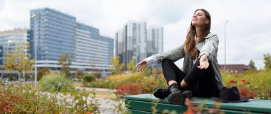 Öğle yemeği molasında genç bir kadın parkta yoga pozisyonunda oturup ofis binalarının arka planında rahatlıyor..