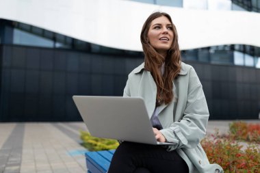 Dizüstü bilgisayarı olan genç bir öğrenci şehrin arka planında bir bankta oturuyor..