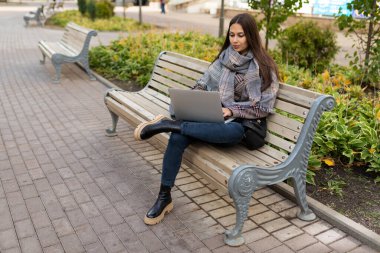 Genç bir kadın internette bilgisayarda çalışıyor. Parkta, üzerinde kalın giysiler var..