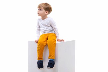 Meraklı bir çocuk beyaz bir kübün üzerinde soldaki beyaz duvara bakıyor.