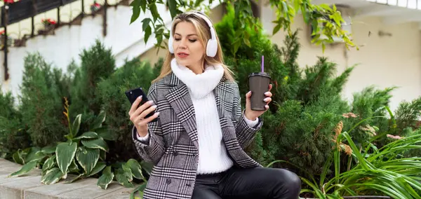an adult woman in headphones chooses a track on a mobile phone with a cup of coffee in her hands.