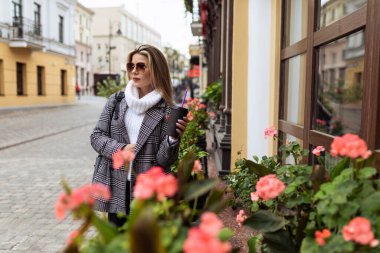 Güneş gözlüklü, sıcak sonbahar elbiseli şık bir yetişkin kadın ve çiçeklerle süslenmiş bir şehir kafesinin yanında gülümseyerek uzaktaki bir yere bak..