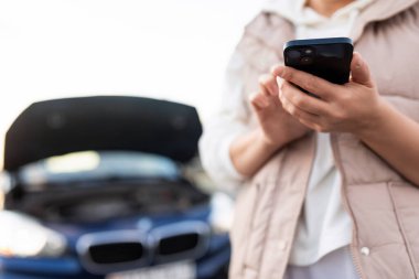 Saha derinliğinde bir fotoğraf, cep telefonuna odaklan, yolda bozuk bir arabanın yanında bir kadın telefonla destek servisini arıyor..