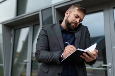 Bir iş adamı, bir ofis binasının arka planında telefonla konuşurken bir not defterine giriş yapar..