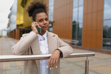 İş kıyafetleri içindeki Afrikalı Amerikalı genç bir kadın bir ofis iş merkezinin arka planında cep telefonuyla konuşuyor..