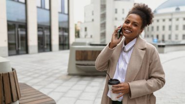 Afro-Amerikan iş kadını elinde bir fincan kahveyle cep telefonuyla konuşuyor..