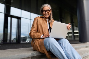 Bir ofis binasının arka planına karşı dizüstü bilgisayarda çalışan kendinden emin yaşlı bir iş kadını..