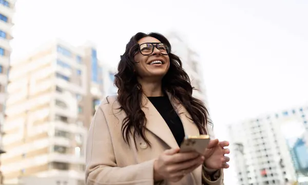 A business brunette with a mobile phone in her hands and glasses for vision against the backdrop of city buildings with a wide smile.