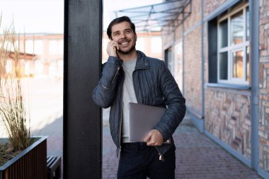 Genç ve başarılı bir işadamı, elinde dizüstü bilgisayarla şehir binalarının arka planında konuşuyor..