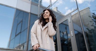 Başarılı iş kadını bir ofis binasının arka planında cep telefonuyla konuşuyor..