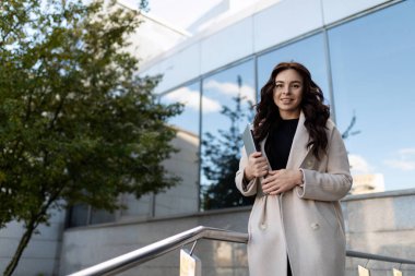 Ofis merkezinin arka planında elinde bir fincan kahveyle başarılı bir modern iş kadınının portresi..