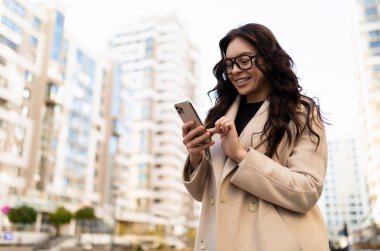 Enerjik modern iş kadını şehir binalarının arka planındaki cep telefonuna bakıyor..
