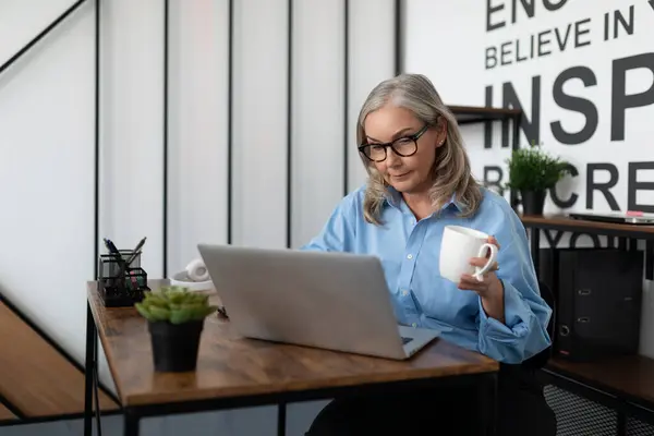 successful woman manager 50 years old works on a laptop with a cup of tea in her hands in the office.