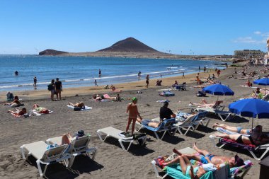 El Medano, Tenerife, İspanya, 8 Mart 2022: Birçok turist, arka planda Kızıl Dağ ile birlikte El Medano 'nun volkanik kumsal plajında güneşlenir ve yıkanır. Tenerife, İspanya