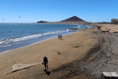 El Medano, Tenerife, İspanya, 8 Mart 2022: İnsanlar arka planda Kızıl Dağ ile El Medano 'nun volkanik kumsal plajı boyunca yürürler. Tenerife, İspanya