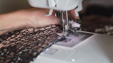 Baskıcı ayak, donanım lambası açık beyaz kumaşa dikiş atarken yakın çekim görüntüsü. Parmakları düzleştiriyor, kararmış arka planda kumaş çiziyor. Sonunda iplik kesiliyor.