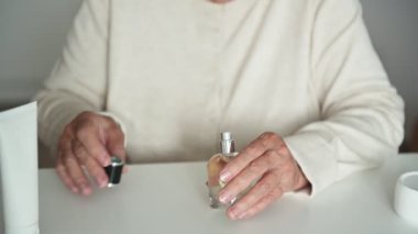 Beyaz bluzlu, buruşuk elleri olan yaşlı kadın masa başında lezzet tatmak için bileğine cam parfüm şişesi ve çikolata serpiştiriyor.