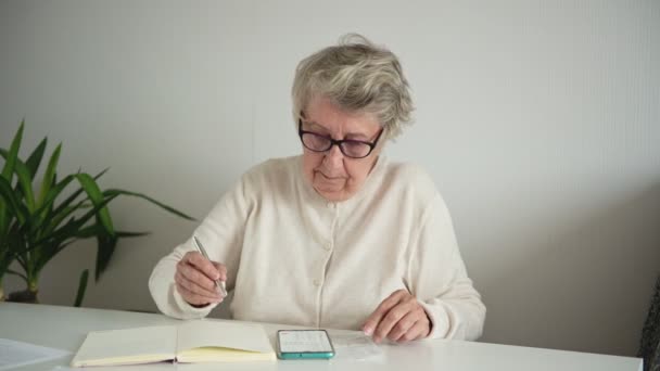 Женщина в очках переписывает расчеты с экрана телефона — стоковое видео