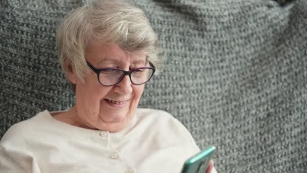 Starsza kobieta patrzy na ekran telefonu uśmiechając się szczęśliwie w domu — Wideo stockowe