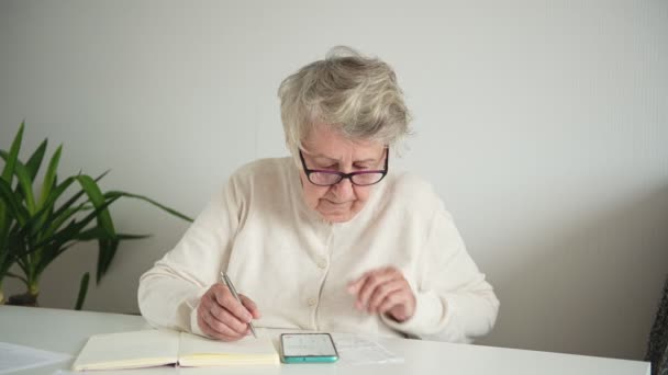Oude vrouw schrijft maandelijkse berekeningen met behulp van smartphone — Stockvideo