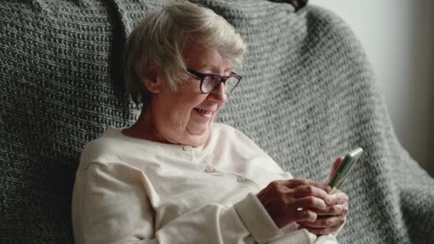 Mulher olha para tela do telefone ri sentado no sofá — Vídeo de Stock