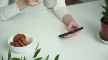 Beyaz ince gömlekli genç bir kadın sosyal medyada akıllı telefon kullanarak ve yulaf ezmeli kurabiyelerle sıcak kahve içerek geziniyor.