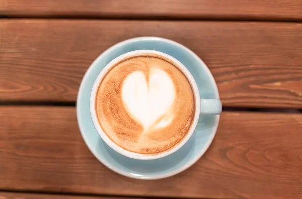 Hati dari busa kopi dalam cangkir biru di atas meja kayu, pemandangan atas. Seni latte. — Stok Foto