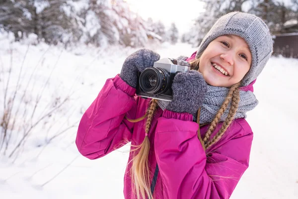 Senyum gadis remaja dengan pakaian hangat, dengan kamera di tangan, terhadap pinus bersalju. Stok Lukisan  