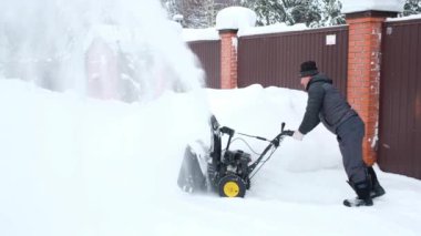 White man removes snow from road with snowblower near fence of suburban house.
