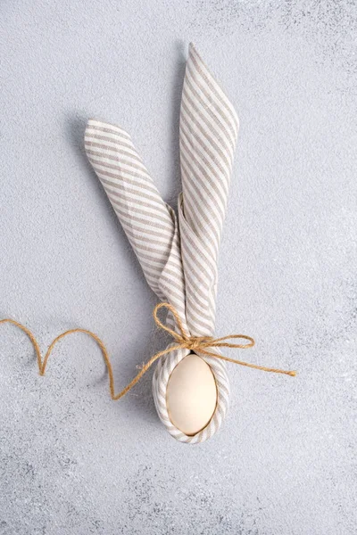 Ovo embrulhado em guardanapo de linho em forma de coelho de Páscoa com orelhas no fundo cinza. — Fotografia de Stock