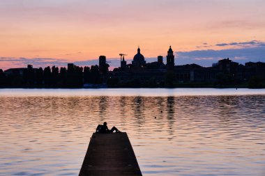 İskelede oturan çift gün batımını seyrediyor ve şehrin ufuk çizgisi, Mantua, İtalya