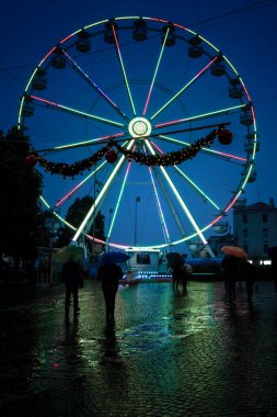 Monza, Italy - December 19 2021: People at night close to the illuminated ferris wheel during the Christmas holidays. Rainy day - People under umbrella