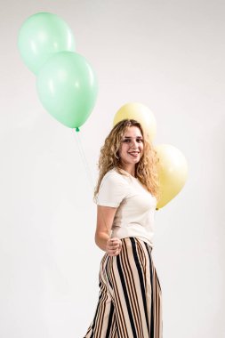 Komik kız, sarışın, açık tişörtlü ve çizgili etekli. Bir kız çıplak ayakla elinde balonlarla beyaz bir arka planda duruyor..
