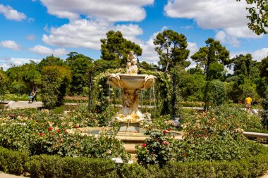 Yazı tipi. Park et. Güller. İspanya 'nın Madrid şehrinde yeşil bitki örtüsü ve çeşitli renklerde çiçeklerle dolu bir parkın yanındaki taştan çeşme. Avrupa. Dikey fotoğrafçılık.