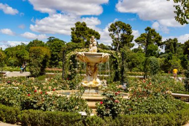 Font. Park. Roses. Stone fountain next to a park full of green vegetation and a variety of flowers of different colors in the city of Madrid, in Spain. Europe. Vertical photography. clipart