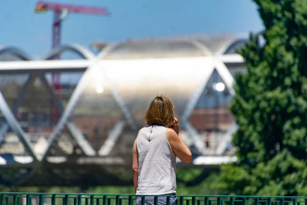 Woman. Person. Middle-aged lady talking on the phone in a park in Madrid. Metal bridge of the Madrid Rio park in the background. Clear day, in Spain. Europe. Horizontal photography.