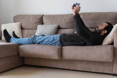 Brown caucasian man with a beard, lying on a sofa while interacting with his blue smart phone
