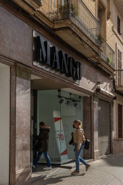 Manacor, İspanya; 20 Ocak 2022: Çok uluslu giyim ve aksesuar şirketi Mango 'nun cephesi, maske takan insanlarla güneşli bir sabah. Yeni normal