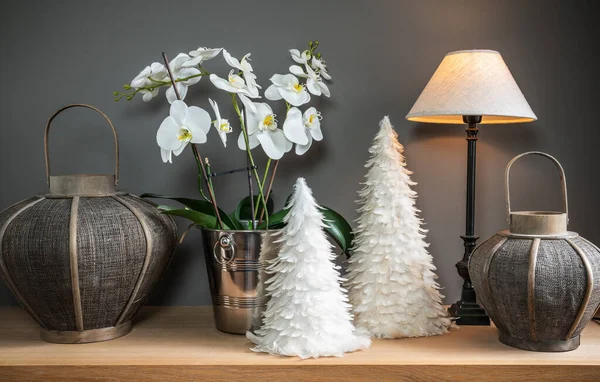 Winter seasonal gifts: White Phalaenopsis flowering plant in silver pot, Christmas trees made of feathers, lanterns and illuminated lamp.