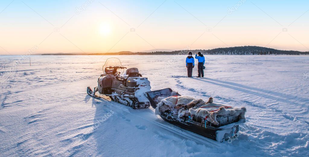 Young couple enjoy sunrise near snowmobile on frozen lake Inari in Finland, Lapland.