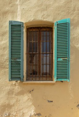 Old window with iron grille and open wooden shutters against yellow outer wall of house in Ajaccio, Corsica.