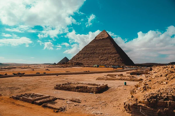 Güneşli bir kış gününde Giza 'nın büyük piramitleri, uzakta birkaç turist otobüsü. Parlak mavi bulutlu gökyüzü, canlı turuncu kum kalıntıları kaplıyor.