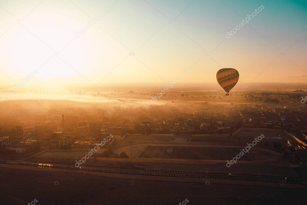 One single hot air balloon in the air, flying low height above the houses. Rearly morning sunrise time, foggy landscape illuminated by sun rays